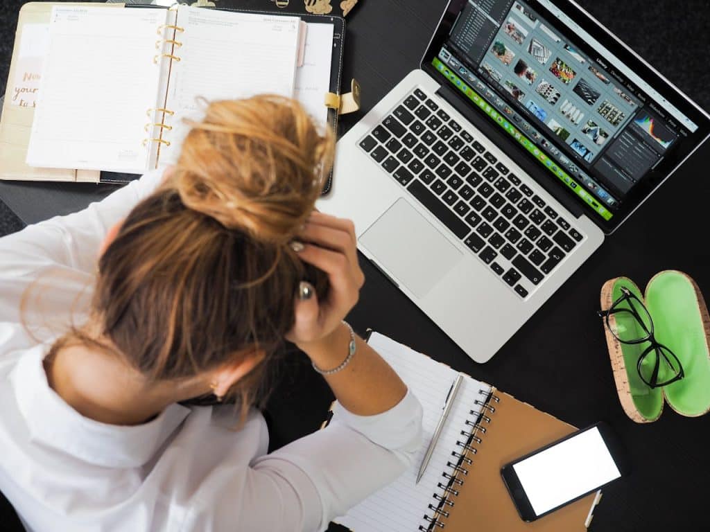 A person is sitting in front of an open notebook and laptop displaying information about their lawn and landscaping company. They have their hands on their head to indicate they are stressed.