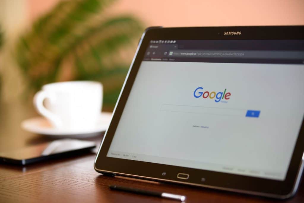 A tablet is sitting on a table with a Google search box displayed with the words "landscaping near me" typed in. There's a cup of coffee and a cellphone next to it.