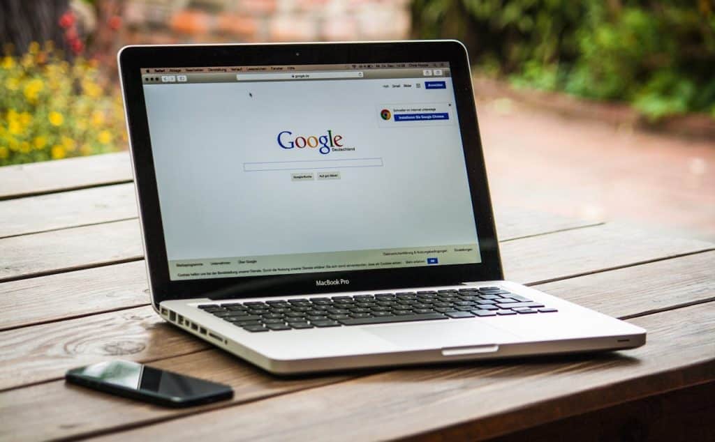 A silver laptop is sitting on a wooden table. The laptop is open and displaying the Google search box with the words "Landscaping near me" typed in.