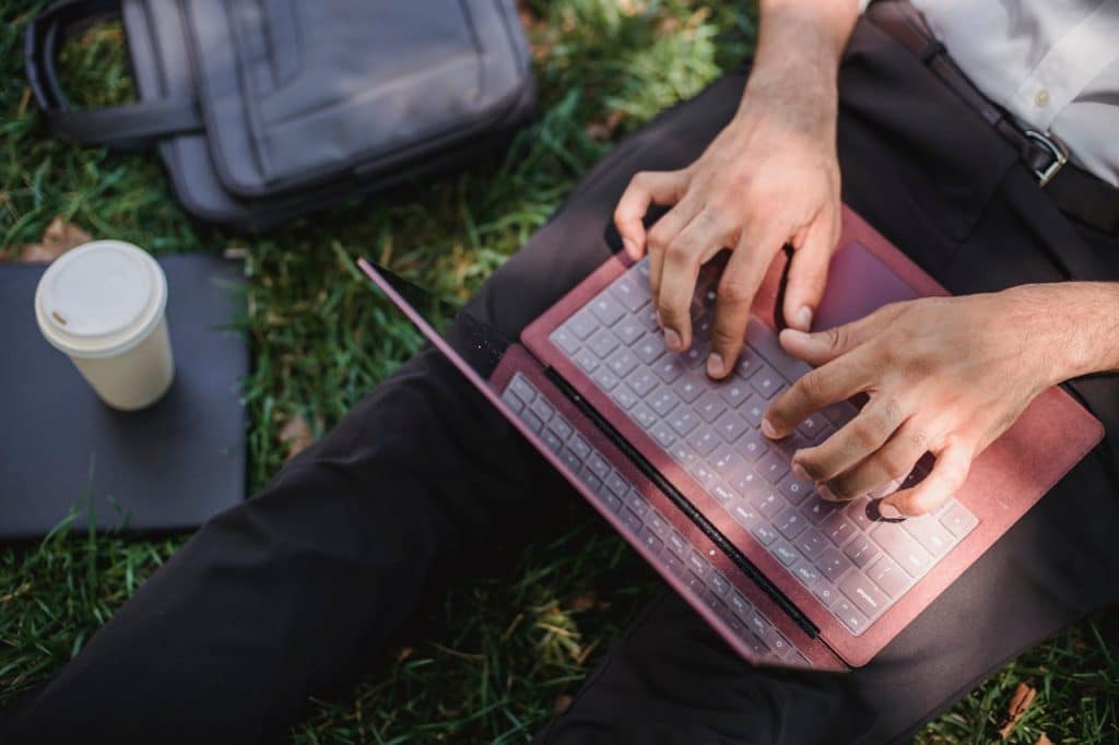 A person is sitting in the grass working on their laptop. They have a briefcase and a cup of coffee next to them.