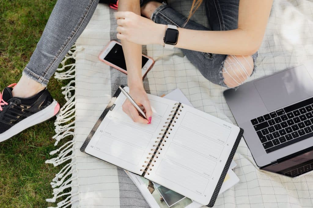 A person is sitting on a blanket in the grass. They have a laptop open next to them while they document jobs in their planner.