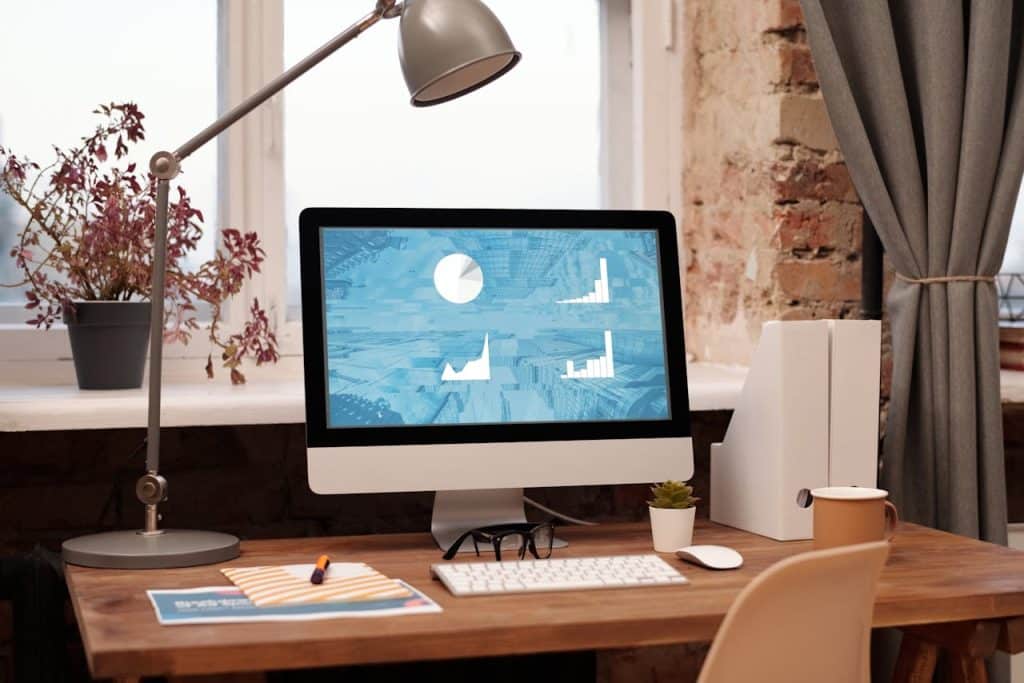 A silver monitor and keyboard sit on a wooden desk in front of a window. The monitor is displaying charts of information on marketing landscaping companies.