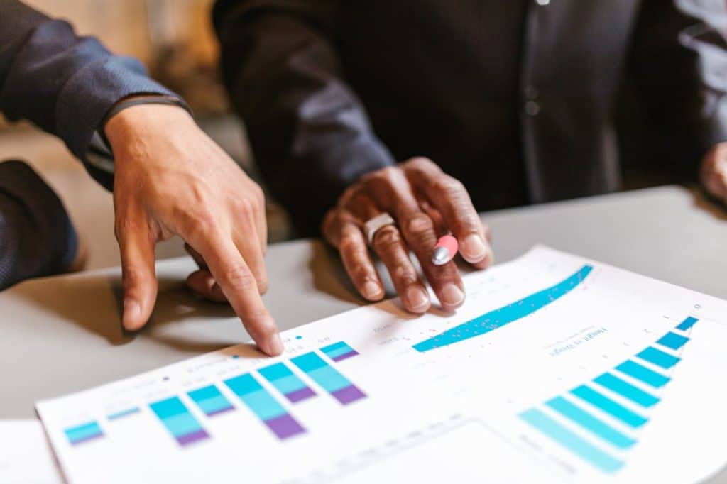 Two landscape business owners analyze a document with bar charts on ROI of landscaping services. One person is holding a pen, the other is pointing at the document.