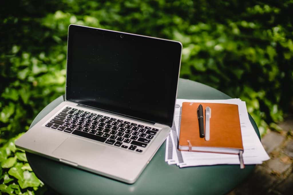 An open laptop, a closed notepad, and information papers on lawn care tips sit on a table outdoors.