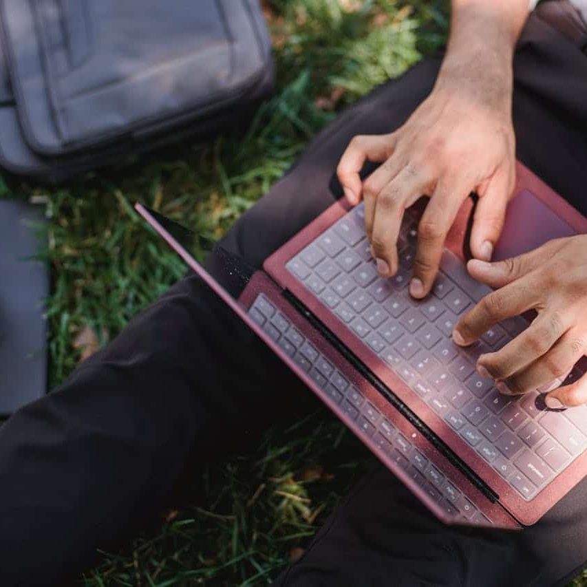A person is sitting in the grass working on their laptop. They have a briefcase and a cup of coffee next to them.
