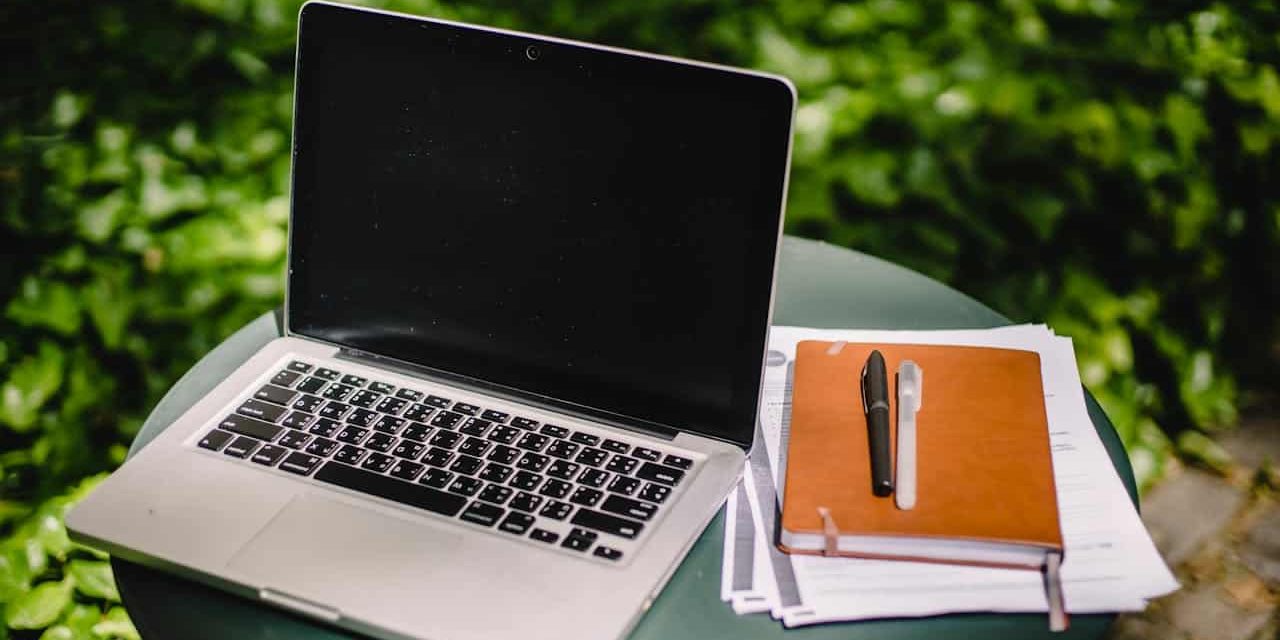 An open laptop, a closed notepad, and information papers on lawn care tips sit on a table outdoors.
